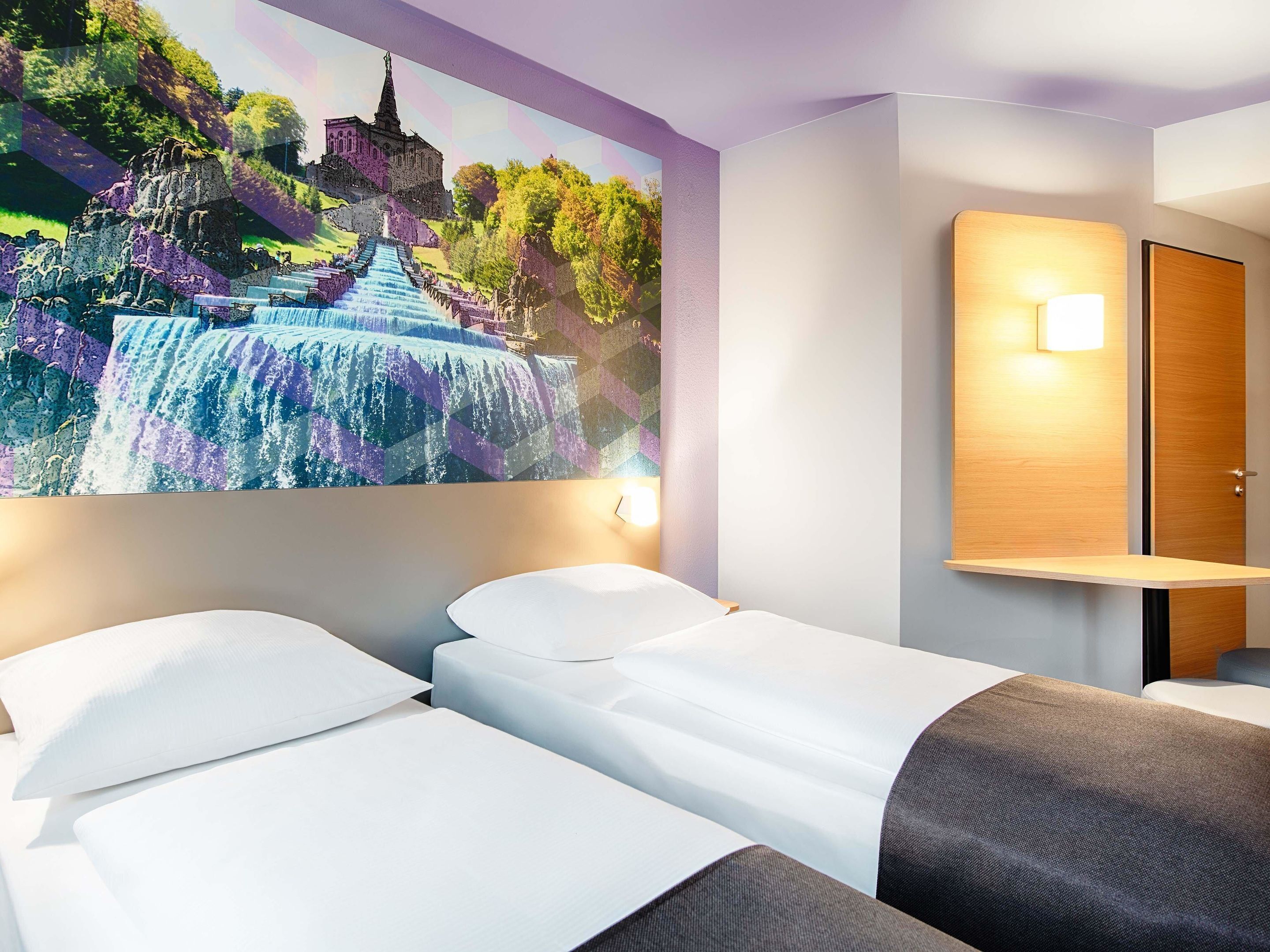 Habitación de hotel con dos camas, un gran mural de una cascada, una mesita de noche de madera y una lámpara fijada a la pared.