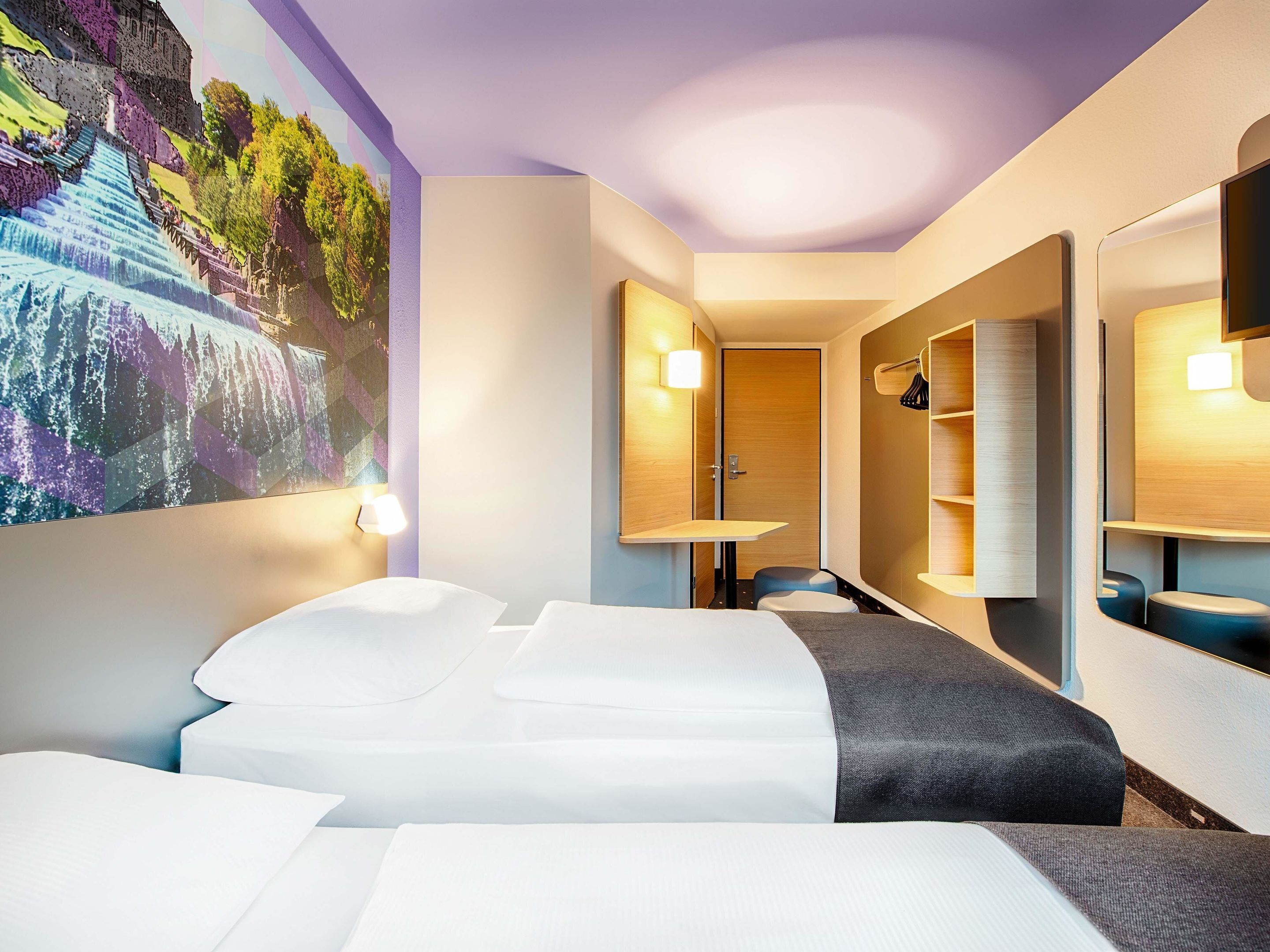 A hotel room with a bed, a desk, a chair, a wardrobe, and a large wall mural.