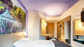 A hotel room with a bed, a desk, a chair, a wardrobe, and a large wall mural. - B&B Hotel Kassel-Süd (Lohfelden)