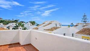 Une terrasse aux murs blancs et aux carreaux de terre cuite, offrant une vue sur une zone côtière avec des bâtiments et des palmiers.
