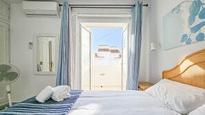 Une chambre d’hôtel avec un lit, un ventilateur, une fenêtre avec des rideaux, une vue sur les bâtiments environnants et un ventilateur de plafond.