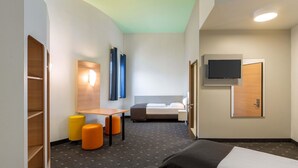 A hotel room with a bed, a desk, two orange ottomans, a TV mounted on the wall, and a wooden wardrobe.