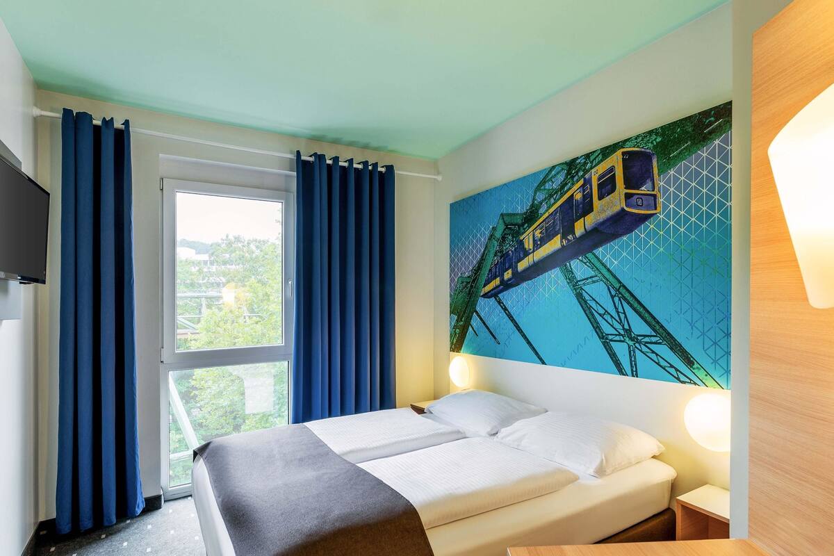 Une chambre d’hôtel avec un lit, une fenêtre avec des rideaux, un tableau représentant un train et une table de chevet.