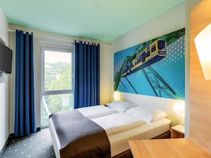 Une chambre d’hôtel avec un lit, une fenêtre avec des rideaux, un tableau représentant un train et une table de chevet.