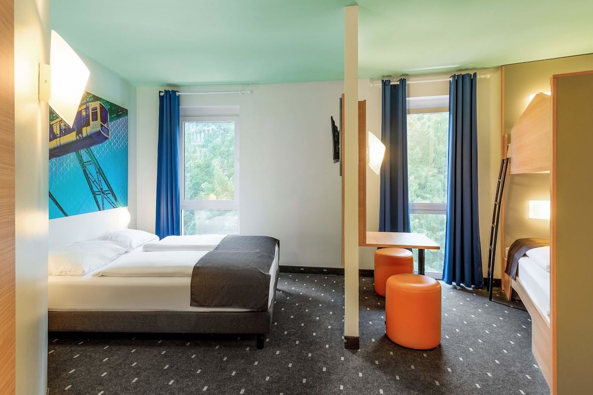 Une chambre d’hôtel avec un lit, deux poufs orange, un bureau et une lampe.