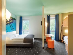 Une chambre d’hôtel avec un lit, deux poufs orange, un bureau et une lampe.