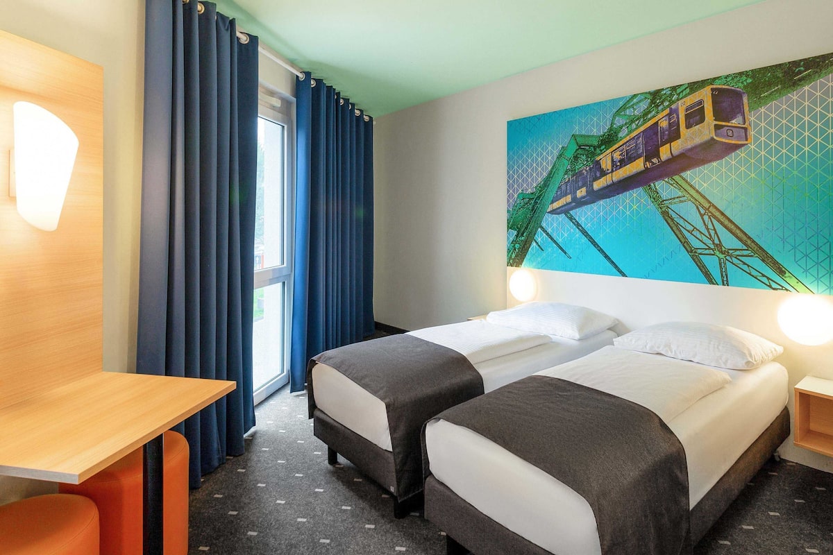Une chambre d’hôtel avec deux lits, un bureau et une grande fenêtre avec des rideaux bleus.