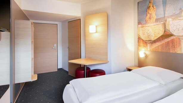 Ein Hotelzimmer mit zwei Betten, einem kleinen Tisch, einem roten Hocker, einer Wandleuchte und einem Bild einer Statue.