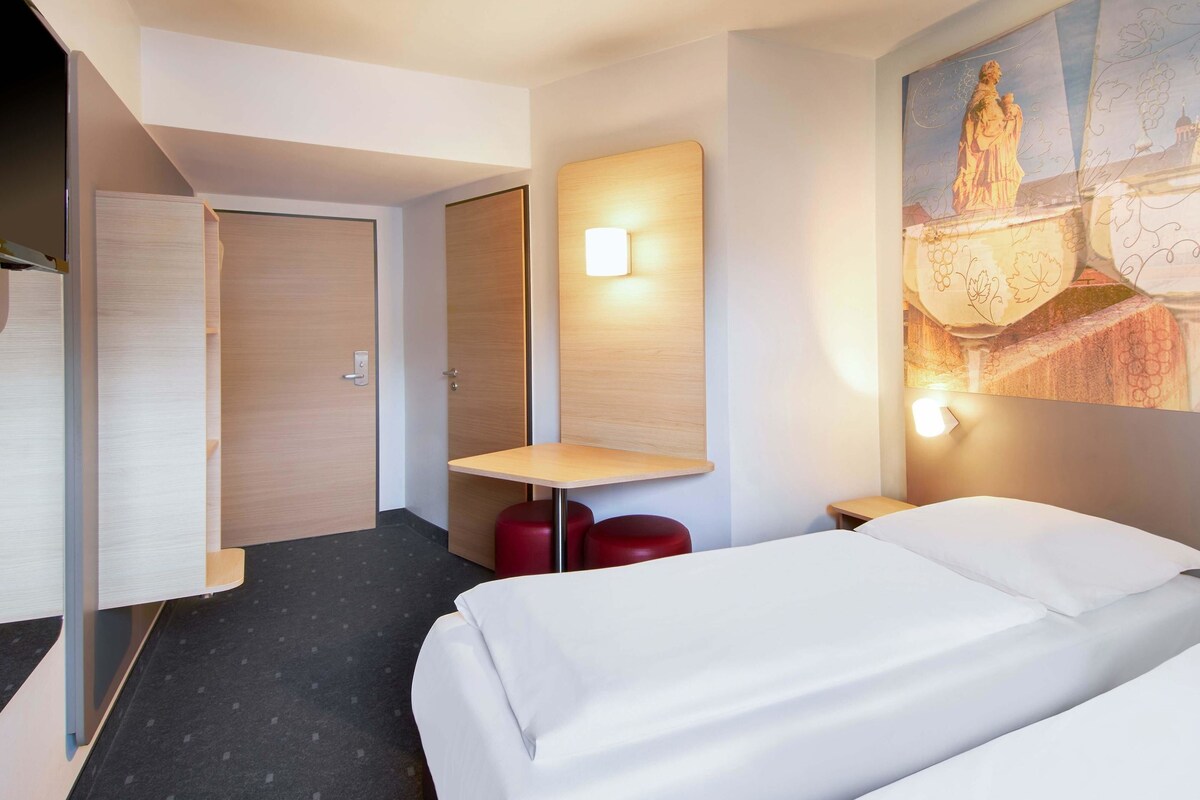 A hotel room with two beds, a small table, a red stool, a wall-mounted light, and a painting of a statue.
