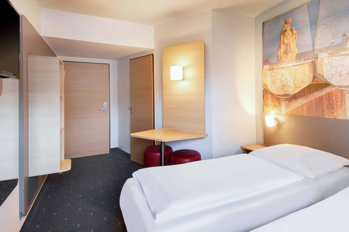 A hotel room with two beds, a small table, a red stool, a wall-mounted light, and a painting of a statue.