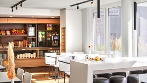 A modern cafe with a central island, white chairs, and a bar area with various beverages and snacks. - B&B HOTEL Würzburg-City (Würzburg)