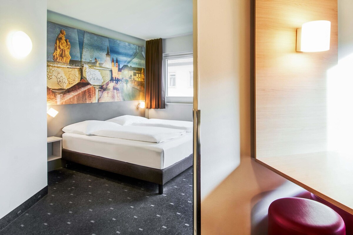 A hotel room with a bed, a painting on the wall, a nightstand, and a wooden headboard with a light fixture.