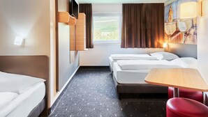 A hotel room with two beds, a small table, a red chair, a window with curtains, and a wall-mounted TV. - B&B HOTEL Würzburg-City (Würzburg)