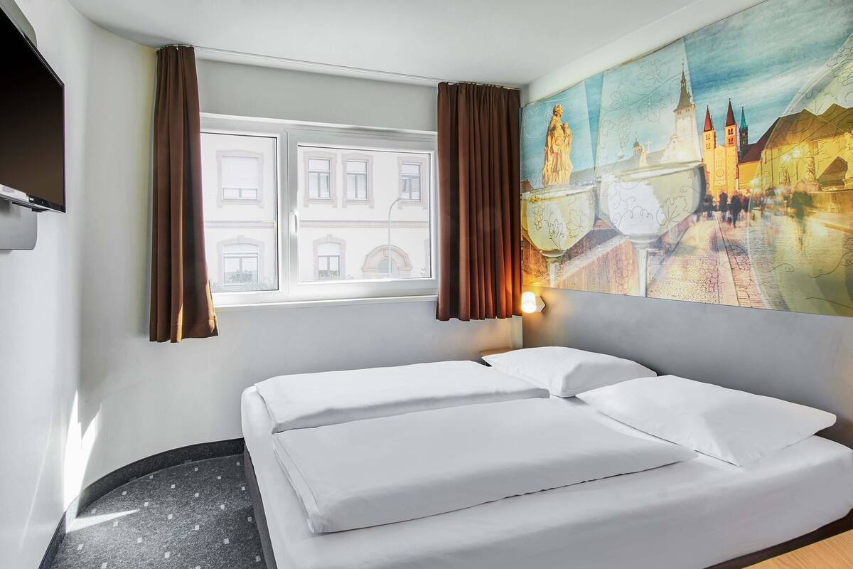 A hotel room with a bed, a window with curtains, a painting of a cityscape, and a wall-mounted TV.