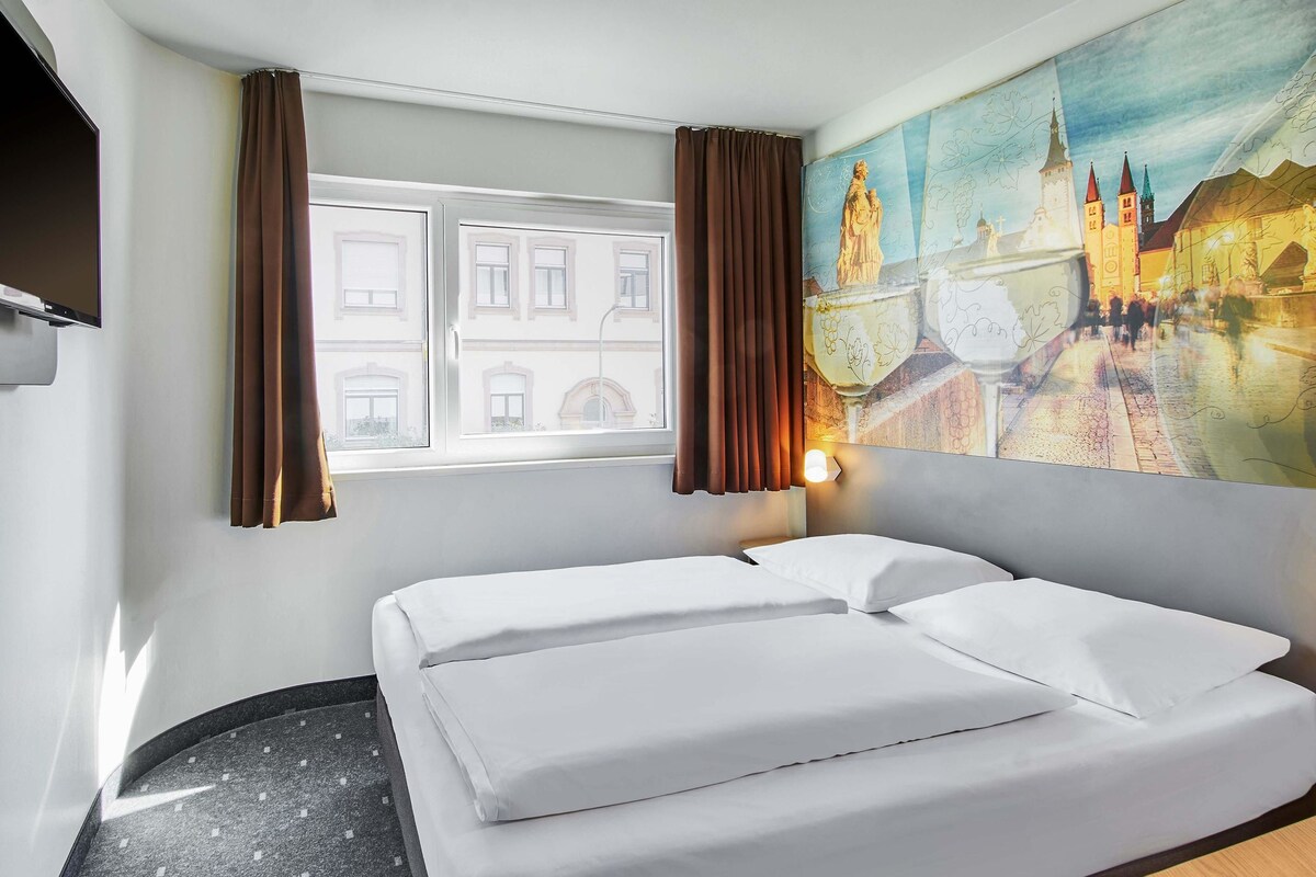 A hotel room with a bed, a window with curtains, a painting of a cityscape, and a wall-mounted TV.