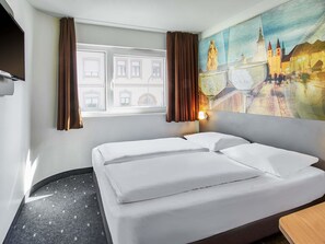 A hotel room with a bed, a window with curtains, a painting of a cityscape, and a wall-mounted TV. - B&B HOTEL Würzburg-City (Würzburg)