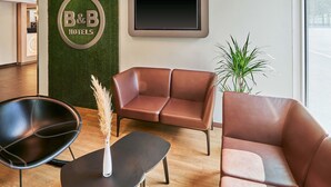 A hotel reception area with a B&B sign, leather sofas, a black chair, a small table, and a potted plant. - B&B HOTEL Würzburg-City (Würzburg)