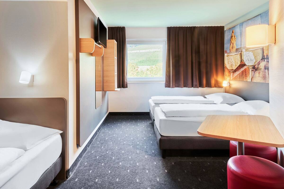 A hotel room with two beds, a small table, a red chair, a window with curtains, and a wall-mounted TV.
