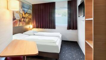Una habitación de hotel con cama, una mesita, televisión y ventana con cortinas.
