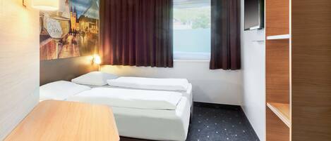 Una habitación de hotel con cama, una mesita, televisión y ventana con cortinas.