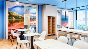 A modern dining area with white tables and chairs, a large window with a view of a lake, and a wall with a scenic painting. - B&B Hotel Kempten (Kempten)