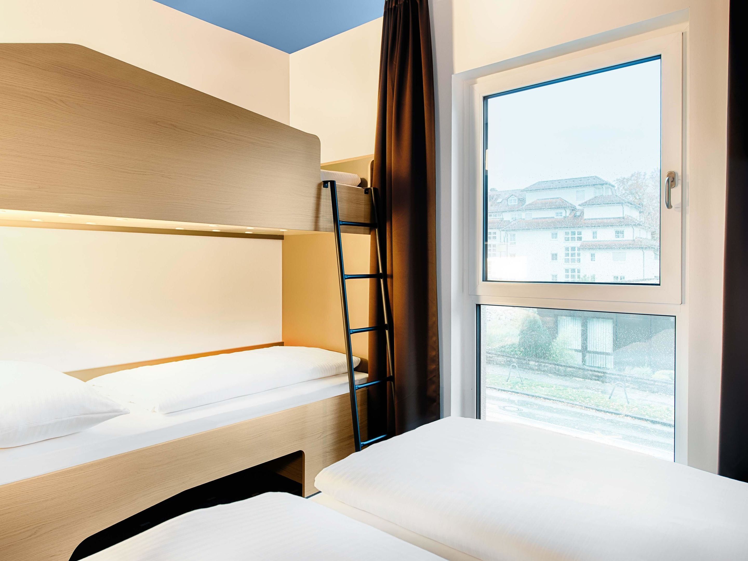 A hotel room with bunk beds, a window with a view of buildings, and a ladder.