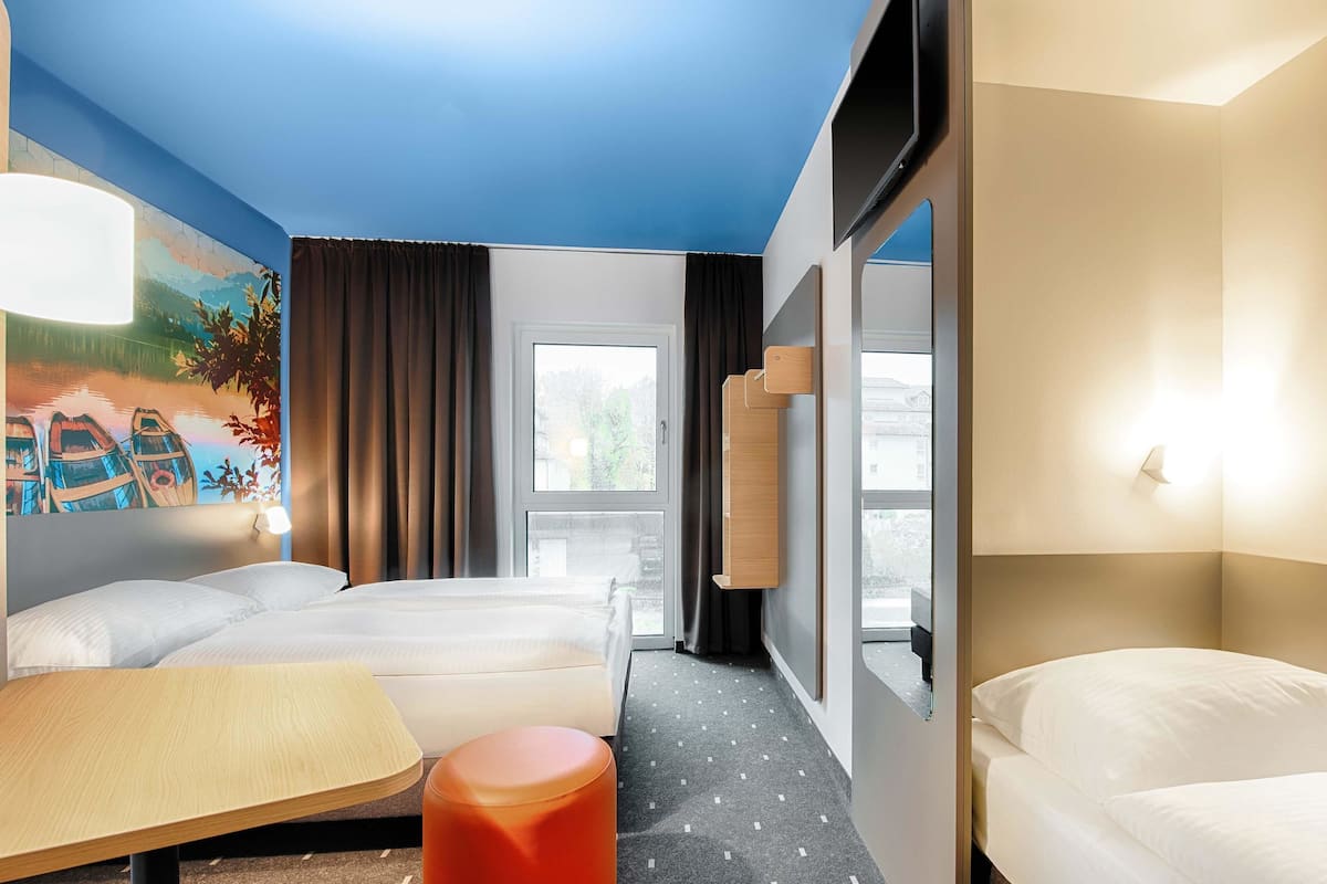 A hotel room with two beds, a wooden table, a red ottoman, a painting of boats, and a view of the outside.