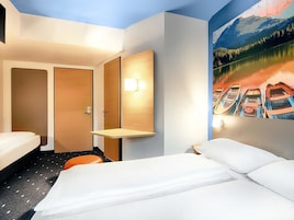 A hotel room with a bed, a small table, a wall-mounted lamp, and a painting of boats on a lake.