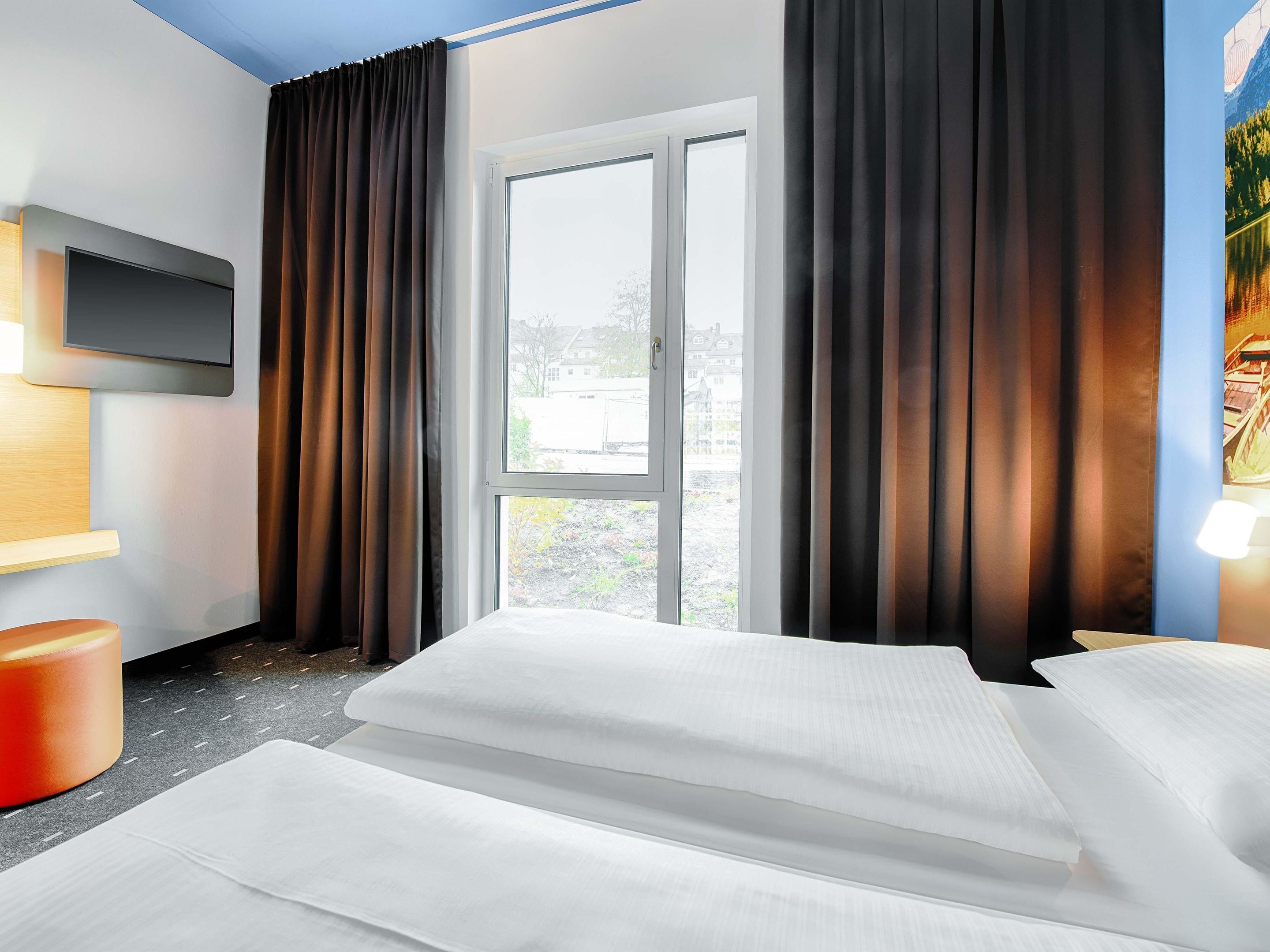 A hotel room with a bed, a TV mounted on the wall, a window with curtains, and a painting on the wall.