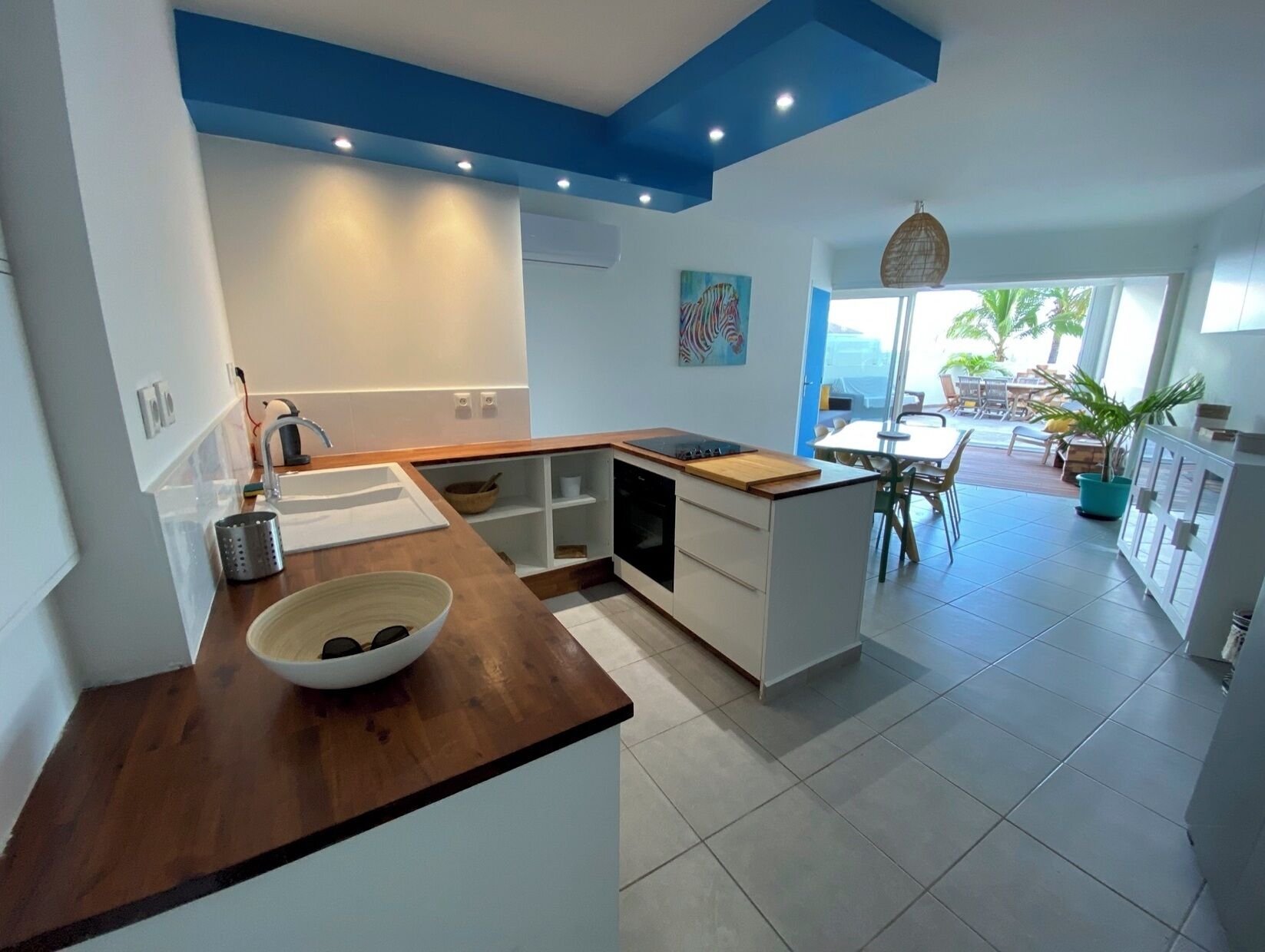 A modern kitchen with a wooden countertop, white cabinets, and a view to a dining area with outdoor seating.