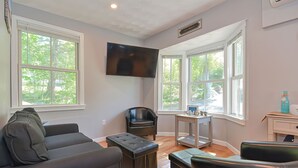 Un salon comprenant un canapé, un pouf et un téléviseur fixé au mur. De grandes fenêtres laissent entrer la lumière naturelle et offrent une vue sur l’extérieur.