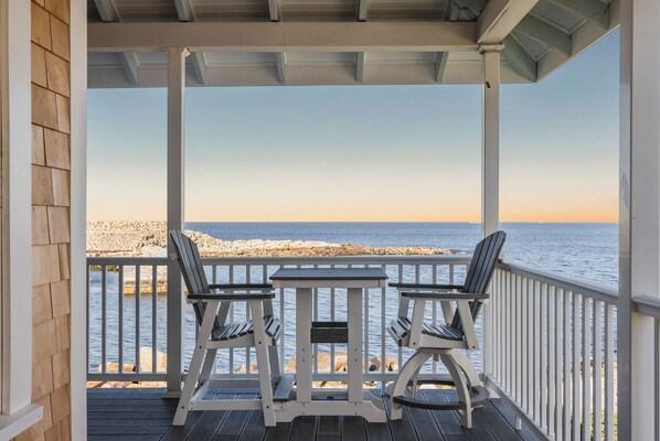 A covered outdoor seating area with two chairs and a table, overlooking a rocky shoreline and the sea.