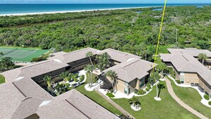 An aerial view of a residential area with multiple buildings, a tennis court, and a beach in the background.