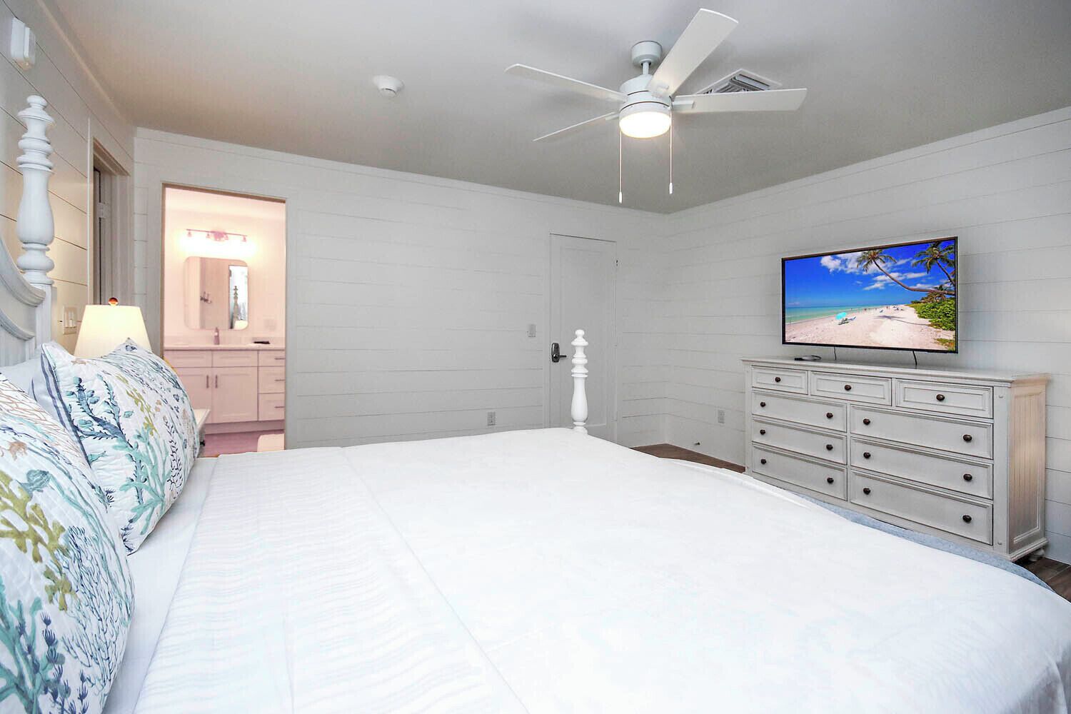 A bedroom with a bed, a dresser, a ceiling fan, and a TV displaying a beach scene.