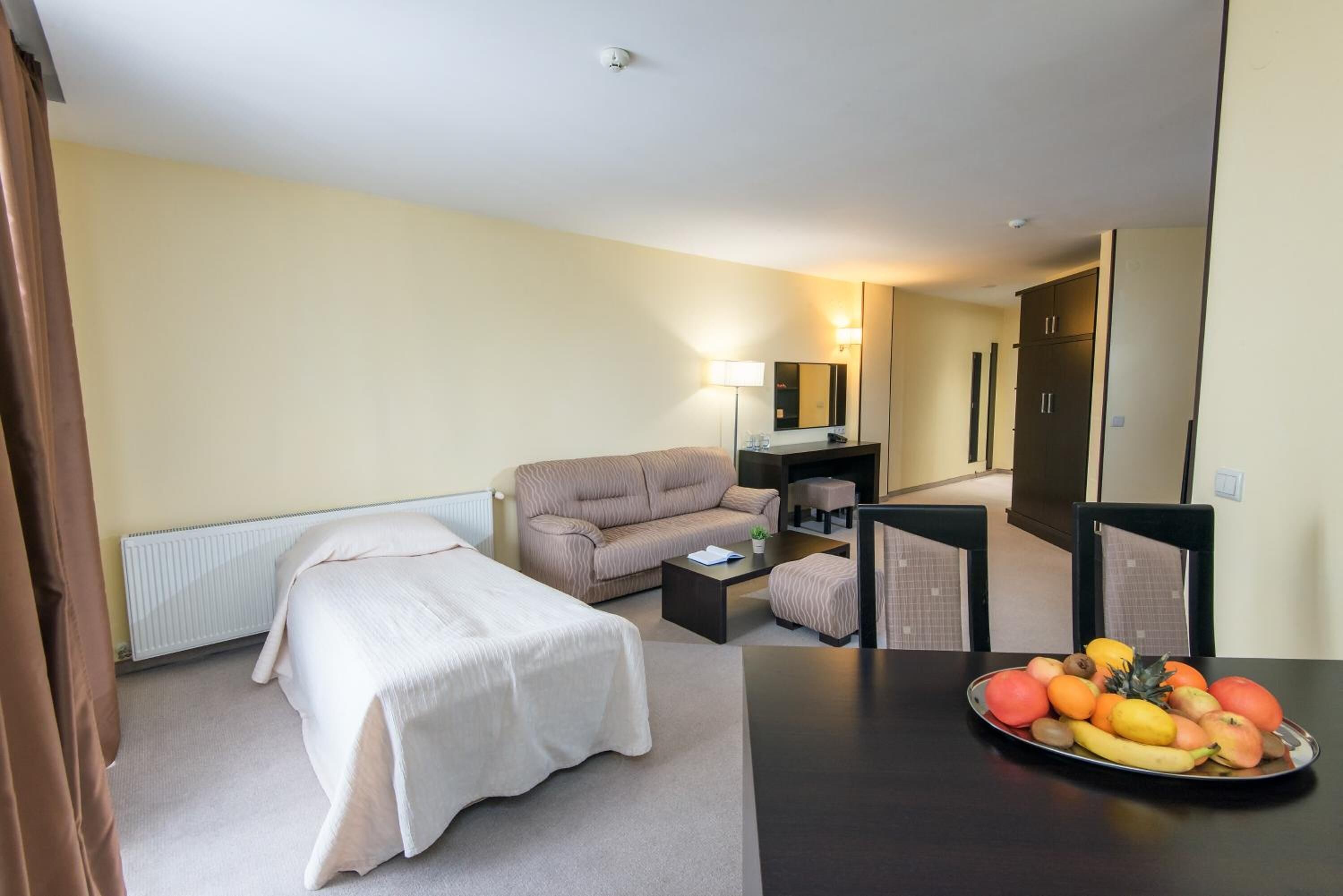 Habitación de hotel con cama, sofá, una mesita, una mesa de comedor con fruta, lámpara y espejo.