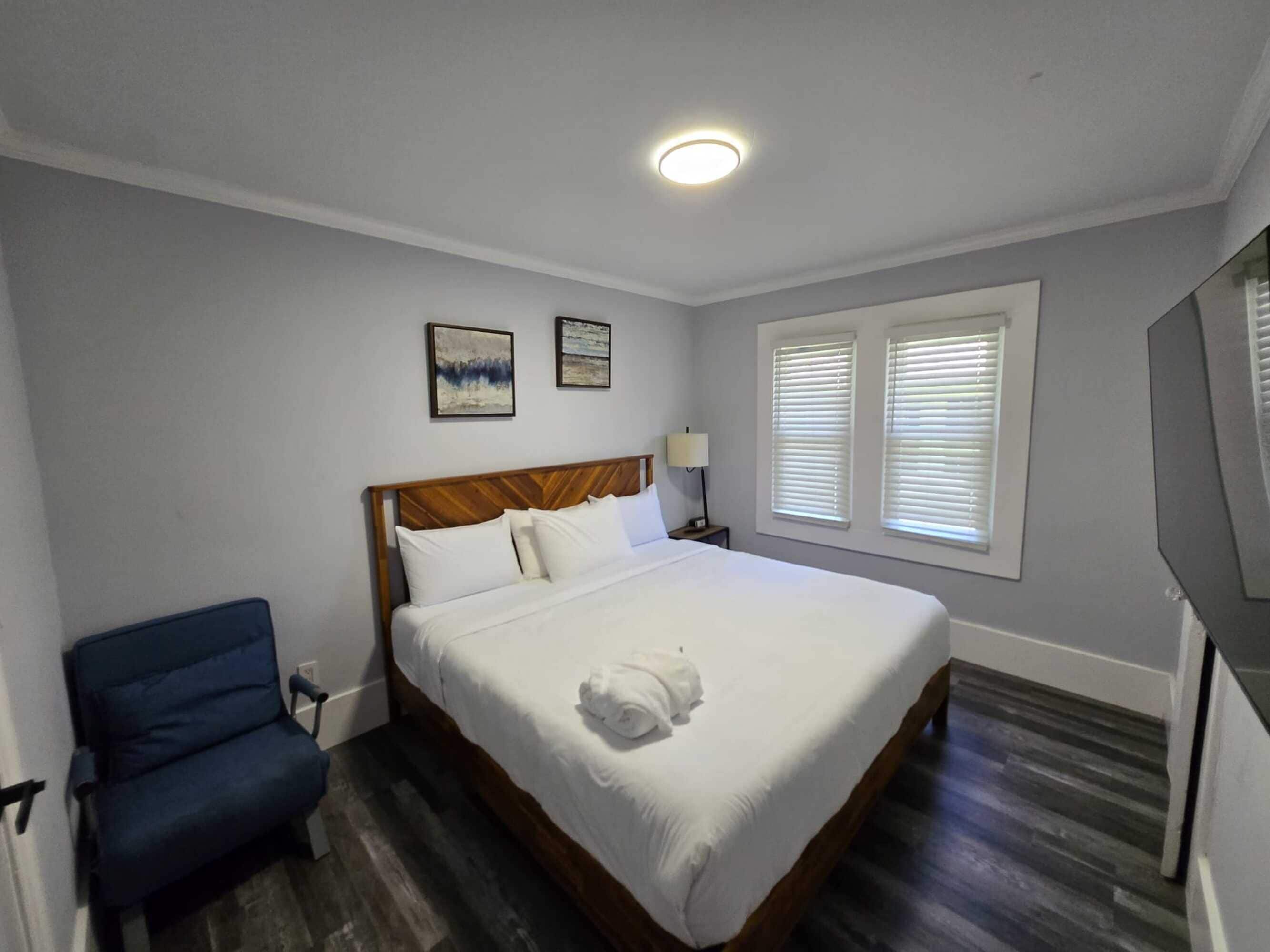 Een net opgemaakt bed met witte lakens en een opgerolde handdoek, een blauwe stoel en een nachtkastje met een lamp.