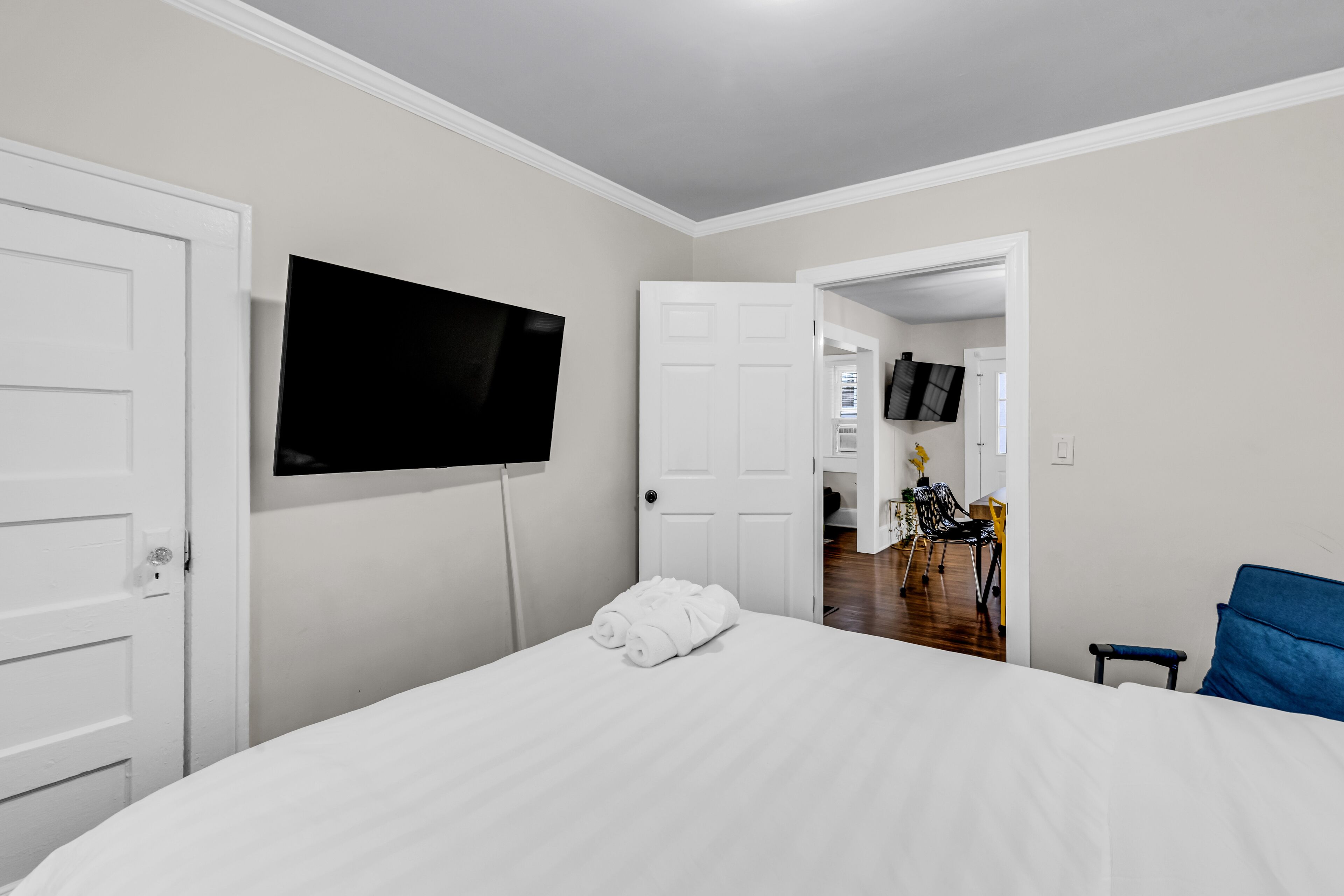 A hotel room with a bed, a TV mounted on the wall, and an open door leading to a dining area.