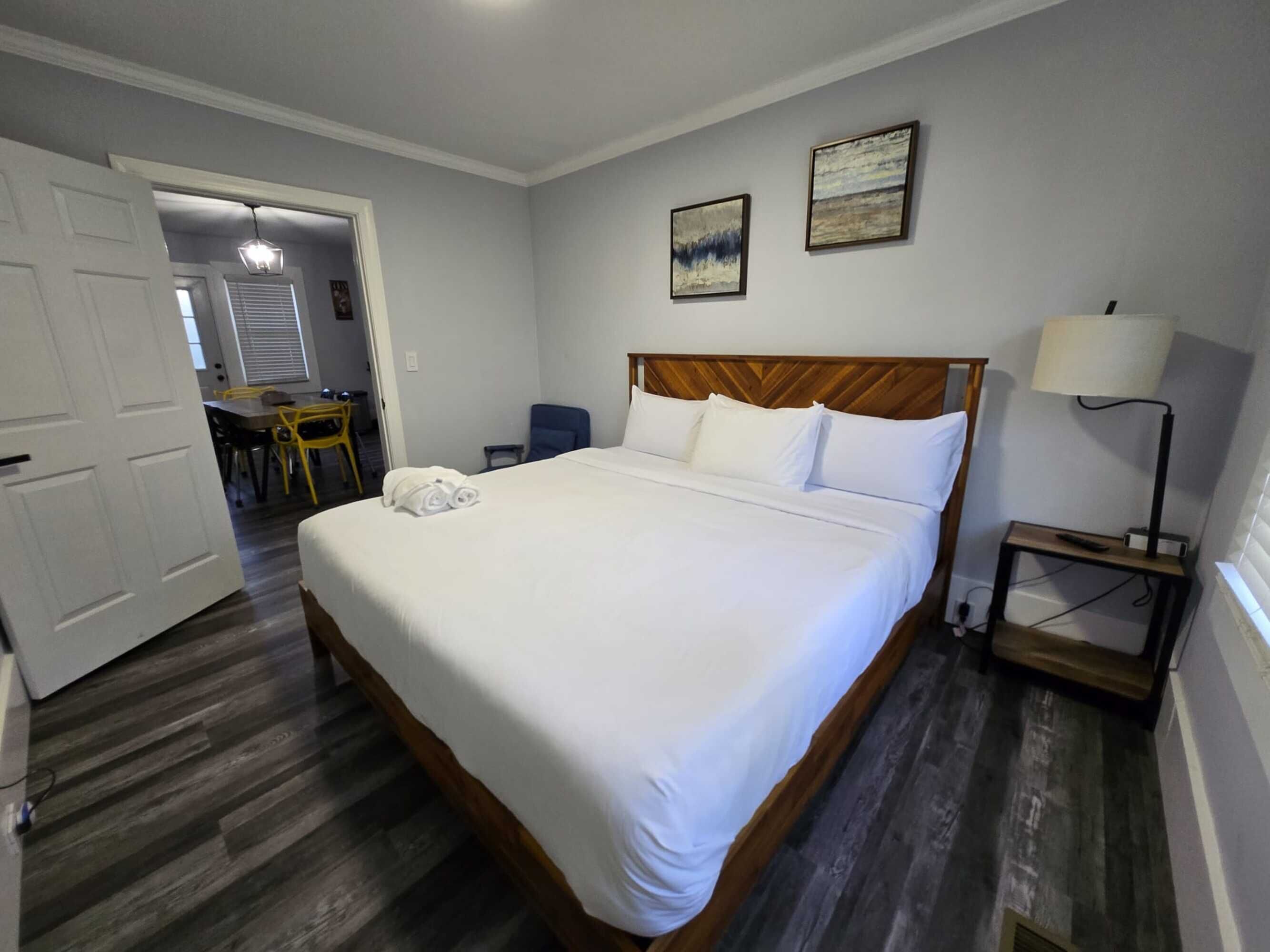 A neatly made bed with white linens, a wooden headboard, and a bedside table with a lamp. There are two paintings on the wall and a chair in the corner.