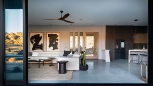 Een moderne woonkamer met een bank, salontafel en kunstwerk. Er is een plafondventilator, een keuken met een koelkast en barstoelen, en een groot raam met uitzicht op rotsen.