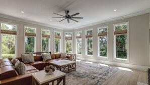 Uma sala de estar ampla com um sofá seccional de couro marrom, uma mesa de centro de madeira e um ventilador de teto. Grandes janelas proporcionam muita luz natural e vista para o verde lá fora.