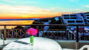A balcony with a table and a vase of flowers, overlooking a coastal town with modern buildings and a sunset. - Xenios Theoxenia Hotel (Aristotelis)