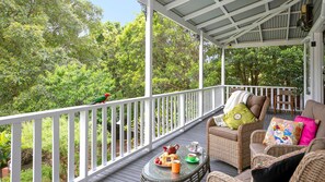 A covered outdoor seating area with wicker furniture, a table with a teapot and pastries, and a bird on the railing. - Lilly Pilly Arthouse– Stay 3 Pay 2 (Stanwell Park)