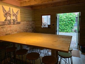 A wooden interior with a large table and stools, a brick wall, and a window with a view of greenery.