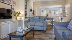Un salon spacieux avec un canapé gris, une table basse et un téléviseur. Au fond, on aperçoit une cuisine avec des meubles blancs et un réfrigérateur.