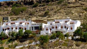 Ein Küstenhaus mit weißen Wänden und roten Ziegeln, gelegen an einem felsigen Hang mit vereinzelter Vegetation.
