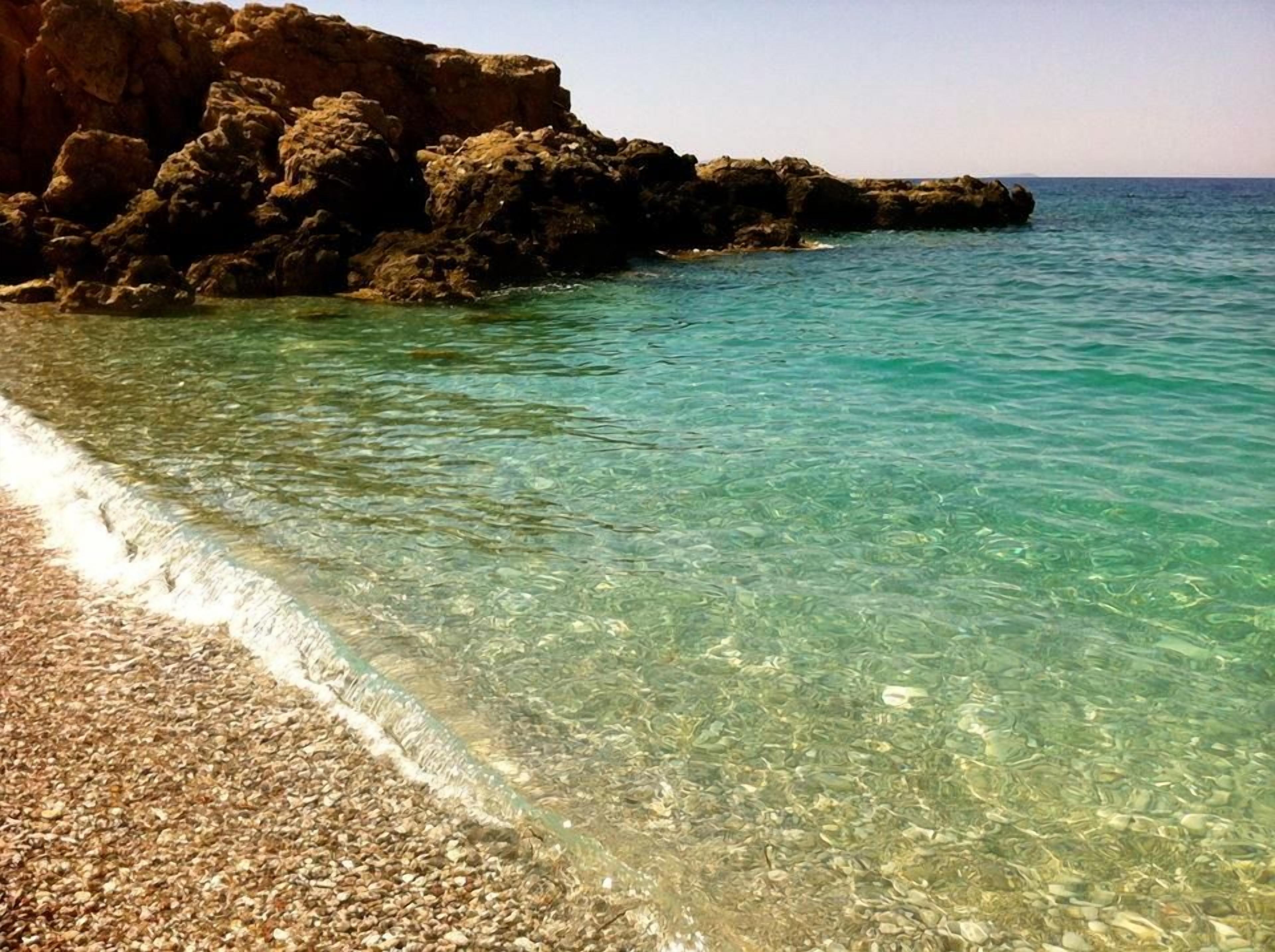 Una playa con agua turquesa cristalina, orilla de guijarros y acantilados rocosos.