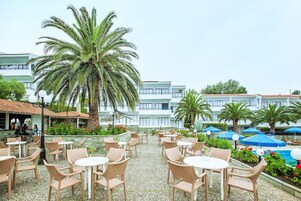 Un espacio al aire libre con sillas y mesas de mimbre, rodeado de palmeras y una piscina.