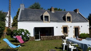 Une maison blanche avec des fondations en pierre, un toit en pente et un petit jardin agrémenté d’une table et de chaises.