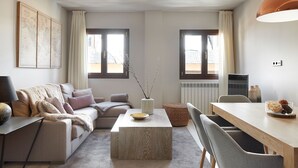 Un salon moderne avec un canapé, une table basse et un coin repas. Il y a deux fenêtres avec des rideaux, un radiateur et une carte du monde accrochée au mur.