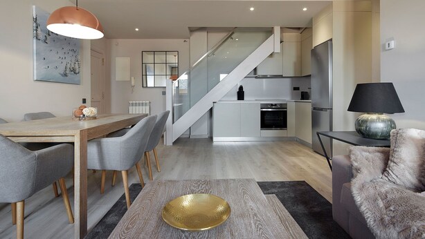 Une cuisine moderne dotée d’un îlot central, d’un coin repas avec une table en bois et des chaises grises, et d’un escalier menant à un niveau supérieur.